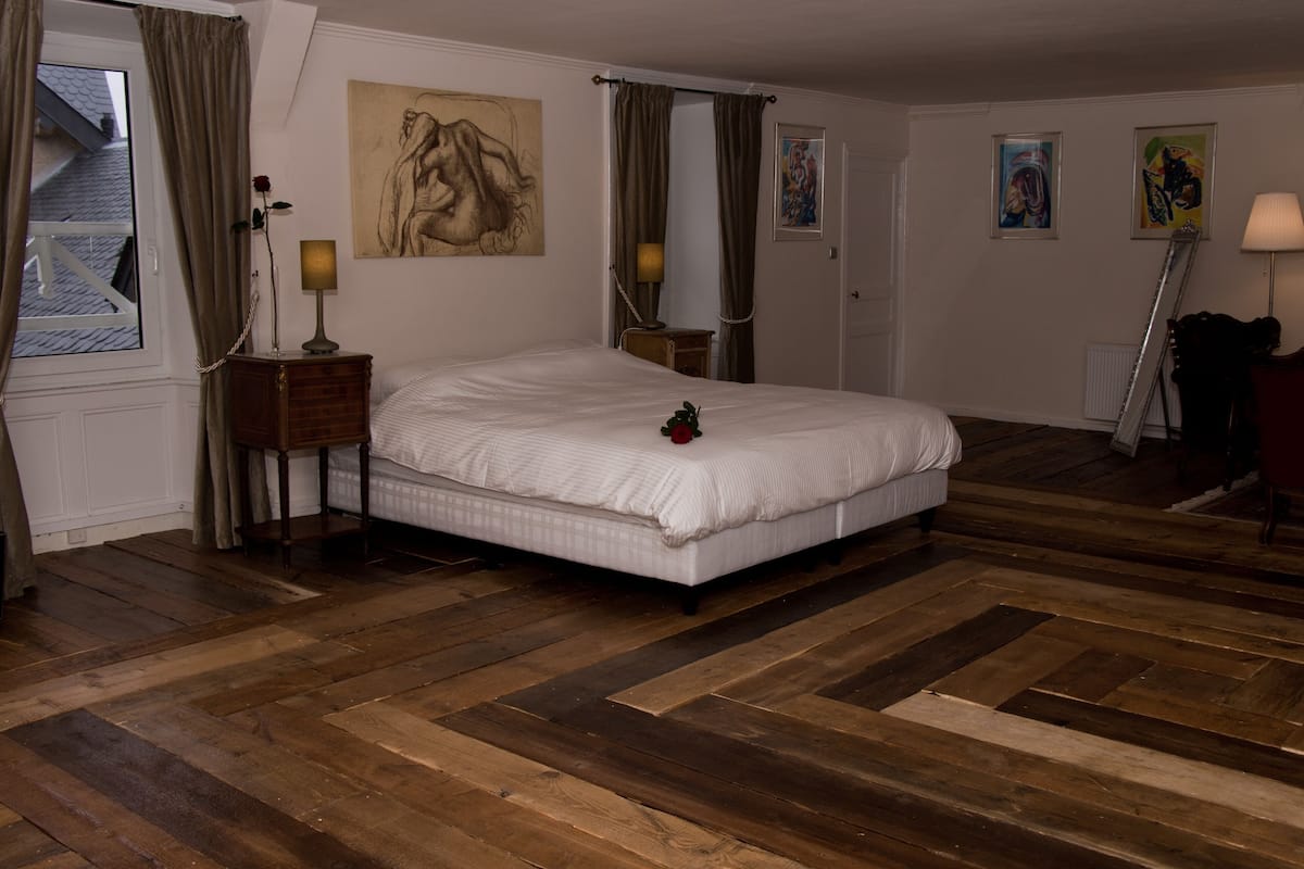 A bedroom with a large bed, wooden flooring, a nightstand with a lamp, and artwork on the walls.