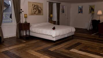 A bedroom with a large bed, wooden flooring, a nightstand with a lamp, and artwork on the walls.