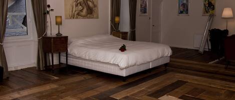Ein Schlafzimmer mit einem großen Bett, Holzdielenboden, einem Nachttisch mit Lampe und Bildern an den Wänden.