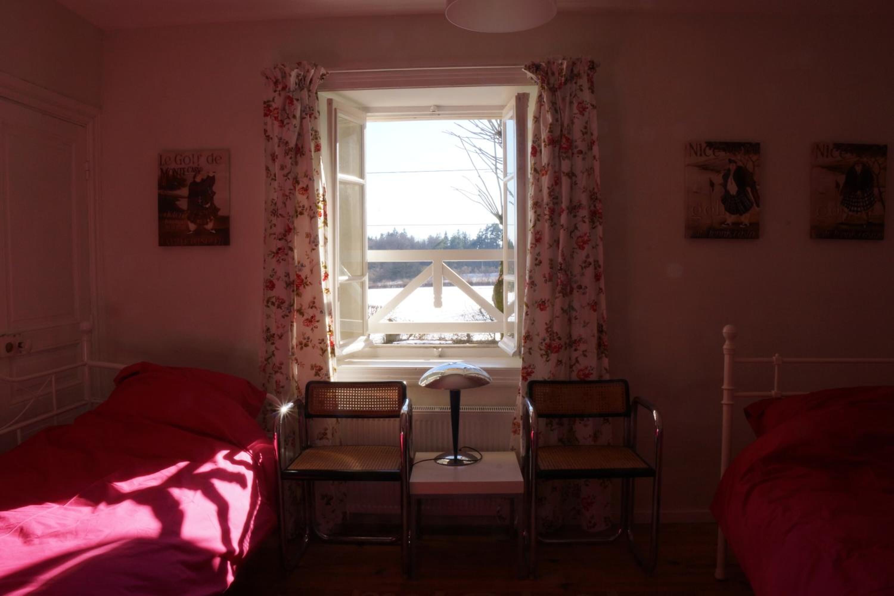 A bedroom with two beds, a window with floral curtains, a table with a lamp, and two chairs.
