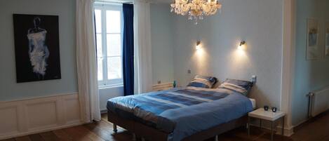 A bedroom with a bed, a chandelier, a window with curtains, and a painting on the wall.