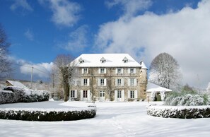 Un edificio grande de piedra con numerosas ventanas y un techo cubierto de nieve, rodeado de arbustos y árboles nevados.