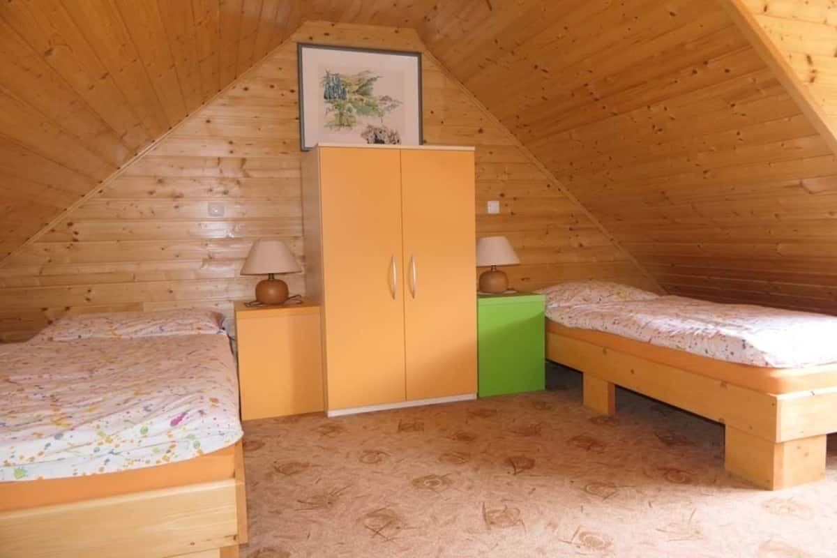 A small, cozy attic bedroom with wooden walls and a sloped ceiling. It includes two single beds, a green nightstand, and a wooden wardrobe.