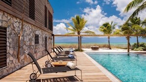 Eine Holzterrasse mit Liegestühlen und einem Pool mit Blick auf einen klaren, blauen Ozean und Palmen.
