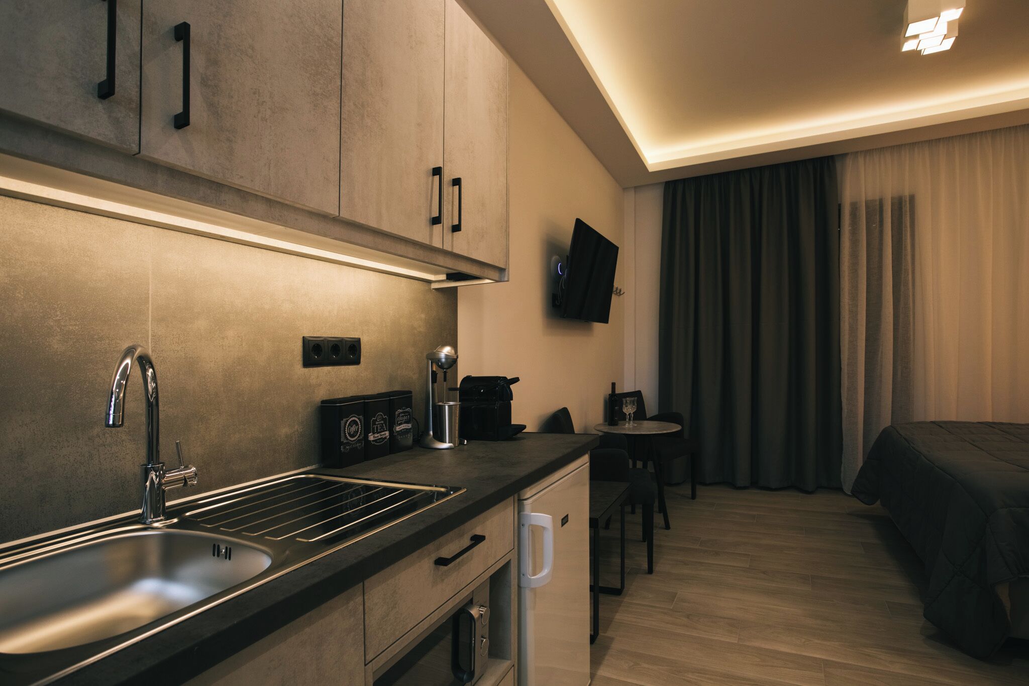 A modern hotel room with a kitchen area, a bed, and a TV mounted on the wall.