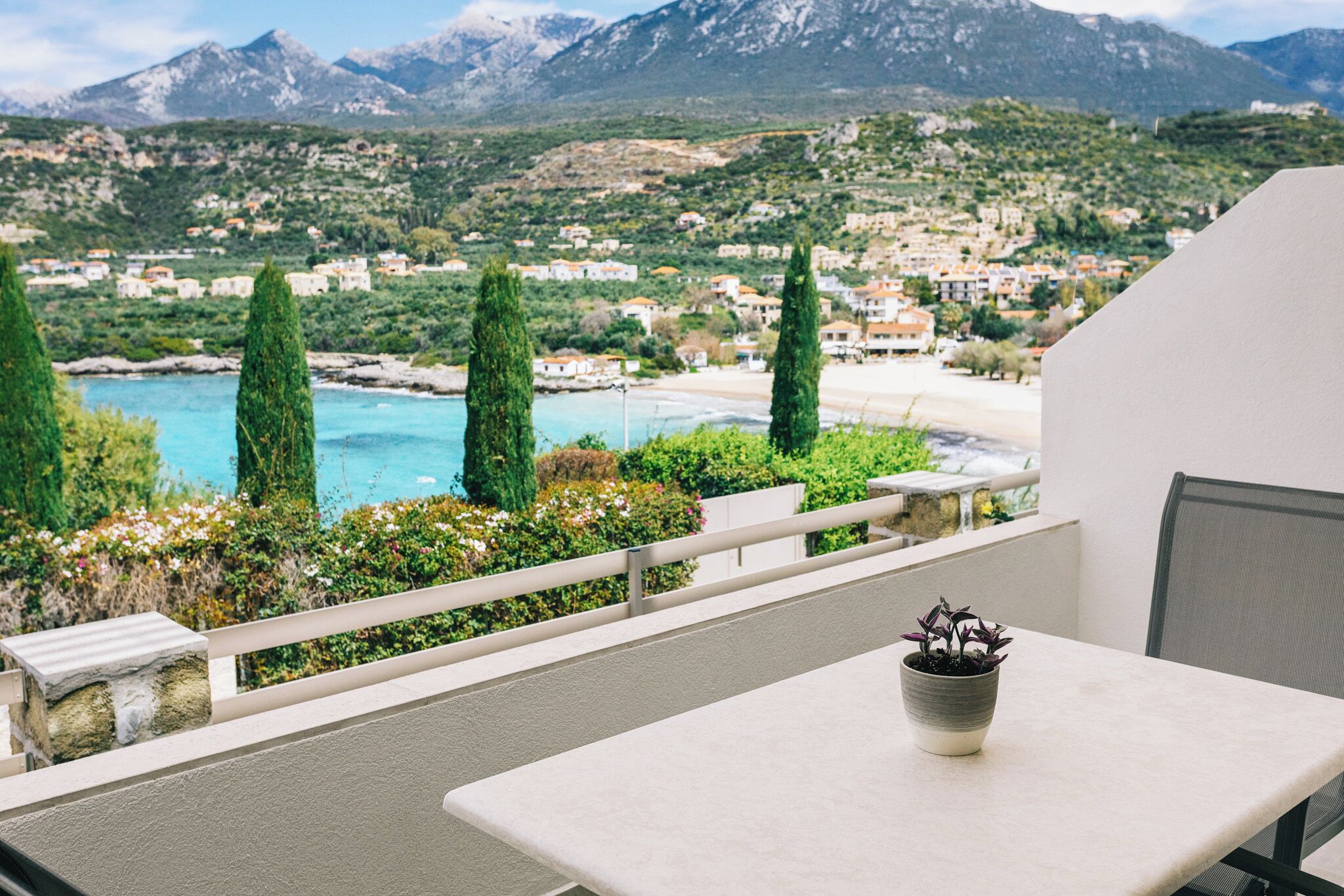 A balcony with a table and a potted plant, overlooking a coastal town and mountains.