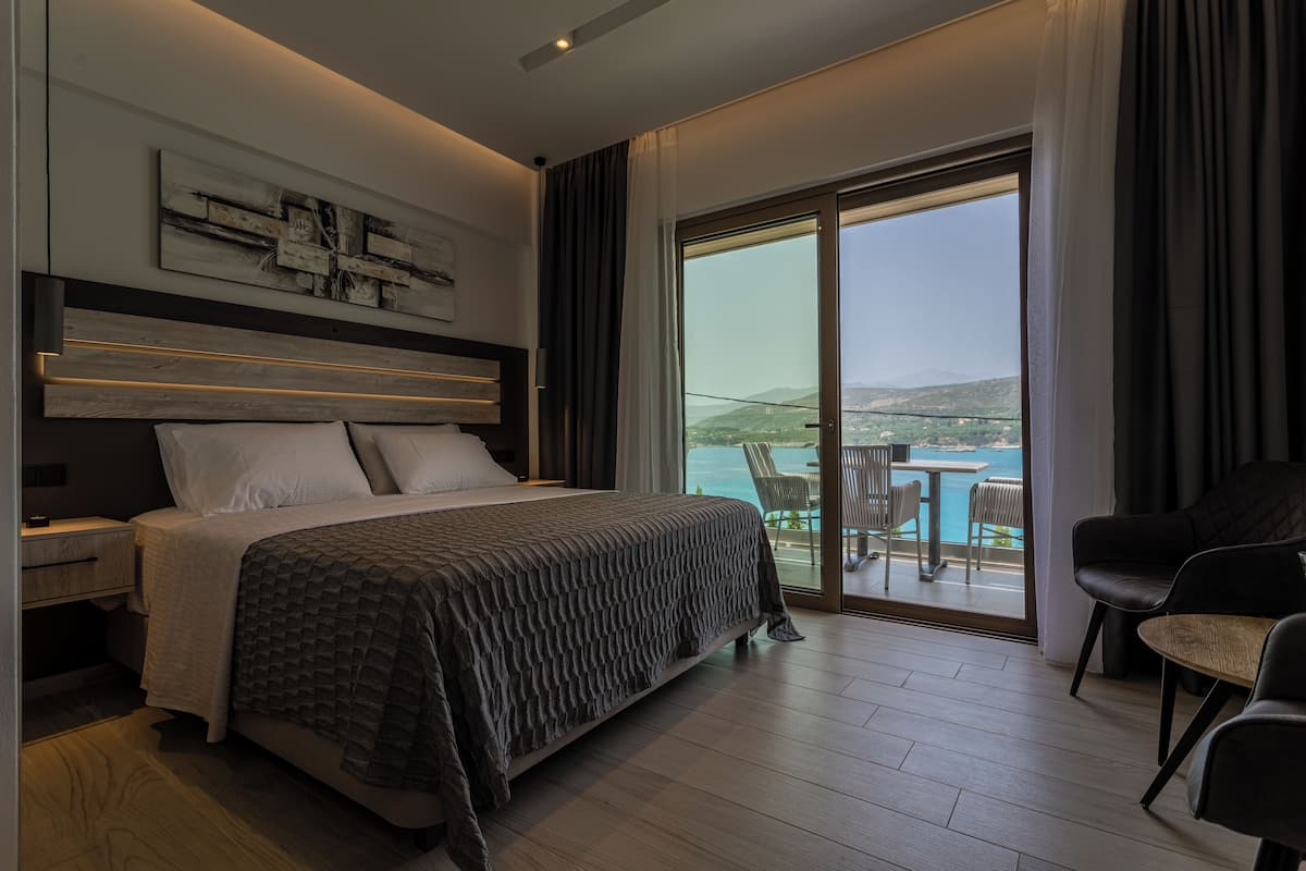 A modern bedroom with a large bed, a headboard with built-in lighting, a view of the sea through a sliding glass door, and a balcony with outdoor seating.