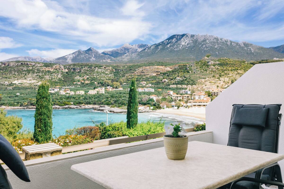 A balcony with a table and chair, overlooking a coastal town with mountains in the background.