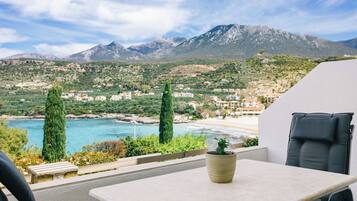 A balcony with a table and chair, overlooking a coastal town with mountains in the background.