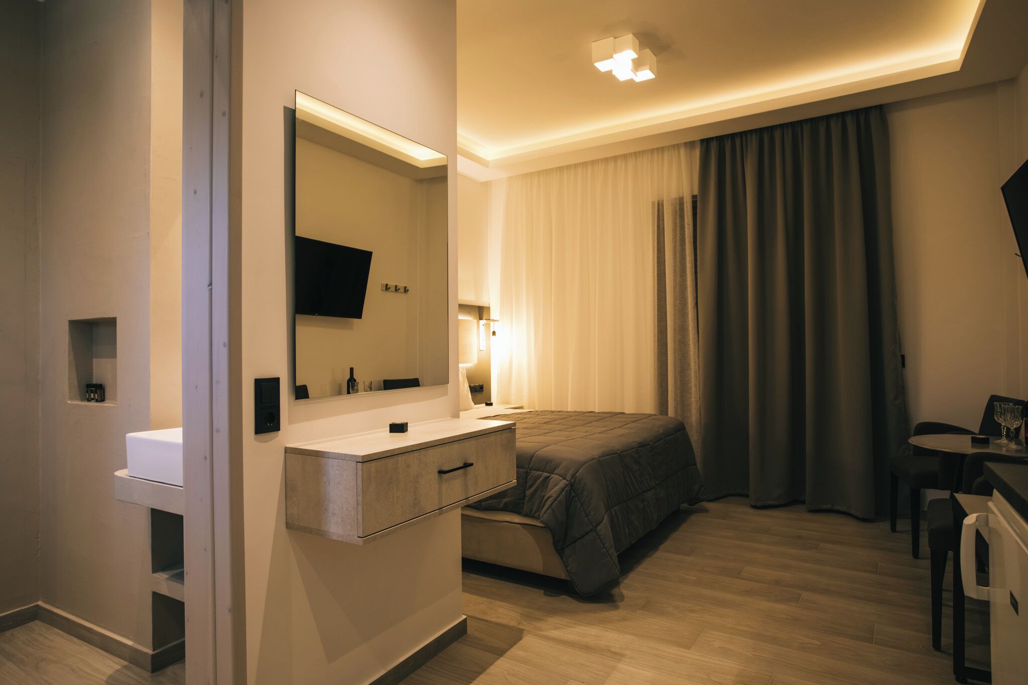 A modern hotel room with a large bed, a bedside table, a flat-screen TV, a desk with a chair, and a window with curtains.