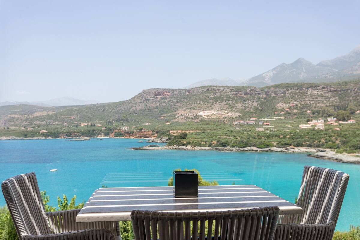 A table with chairs facing a scenic view of a turquoise sea and mountains.