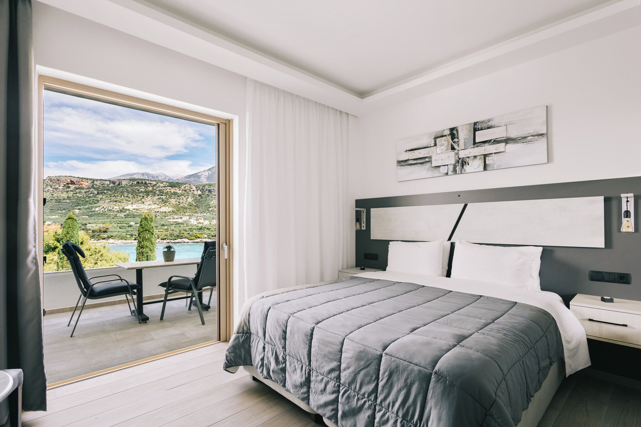 Una habitación de hotel moderna con una cama grande, un balcón con mesa y sillas, y una vista panorámica de las montañas.