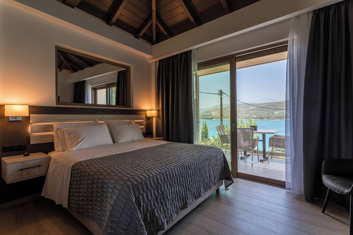A bedroom with a large bed, a nightstand, a mirror, and a view of a balcony with outdoor furniture.