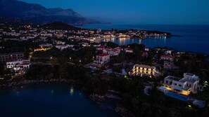 Un pueblo costero al anochecer con edificios a lo largo del paseo marítimo, una iglesia destacada y una montaña en el fondo.