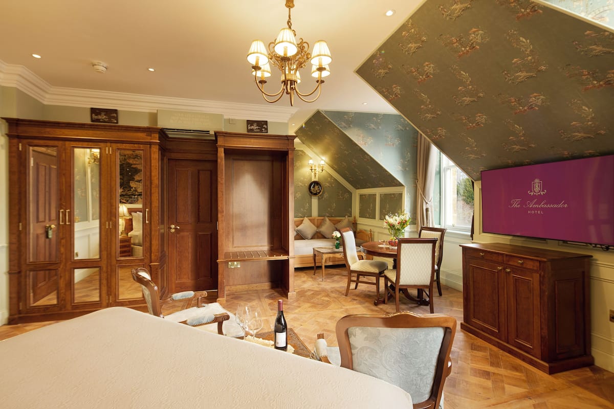 A spacious hotel room with a large bed, a dining area, a seating area, and a view of the outdoors.