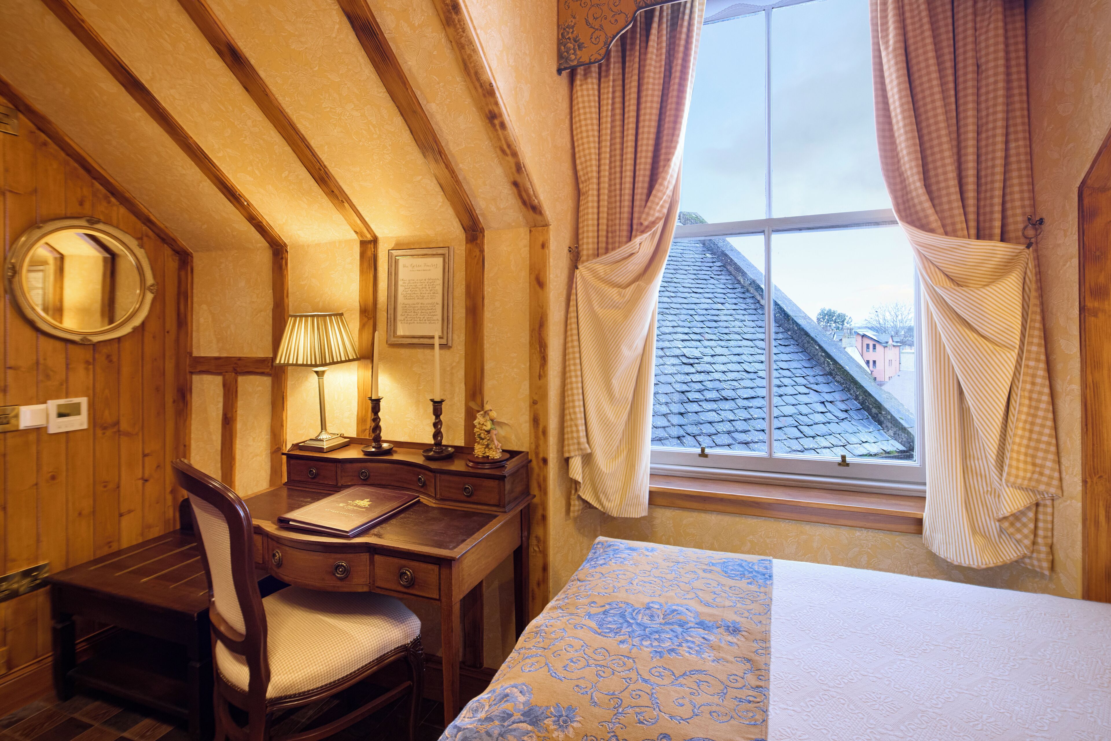 A room with a wooden desk, chair, and a bed with a patterned bedspread. There is a window with curtains and a view of the roof.