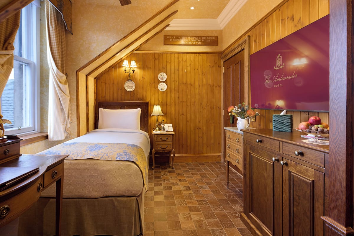 A hotel room with a wooden floor, a bed with a white comforter, a wooden dresser, a nightstand with a lamp, and a sign on the wall.