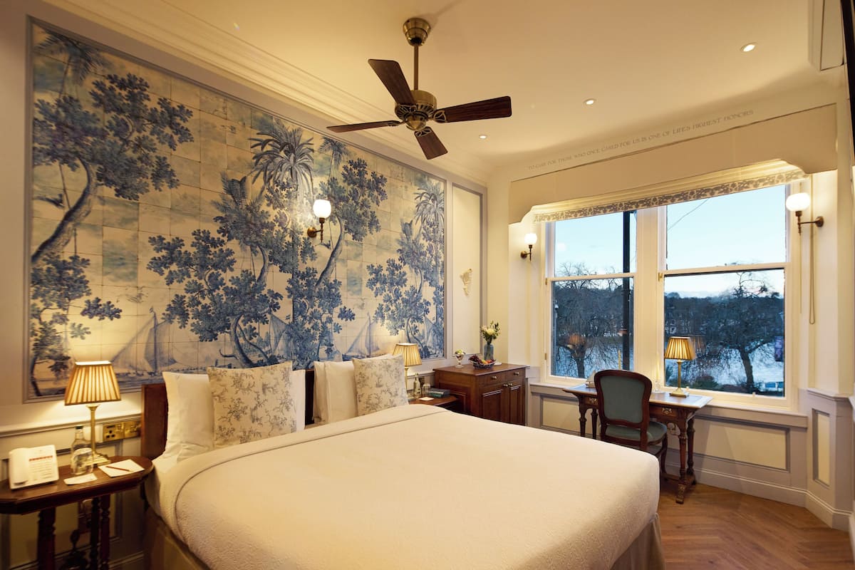 A hotel room with a large bed, a desk, a chair, a ceiling fan, and a window with a view of trees.