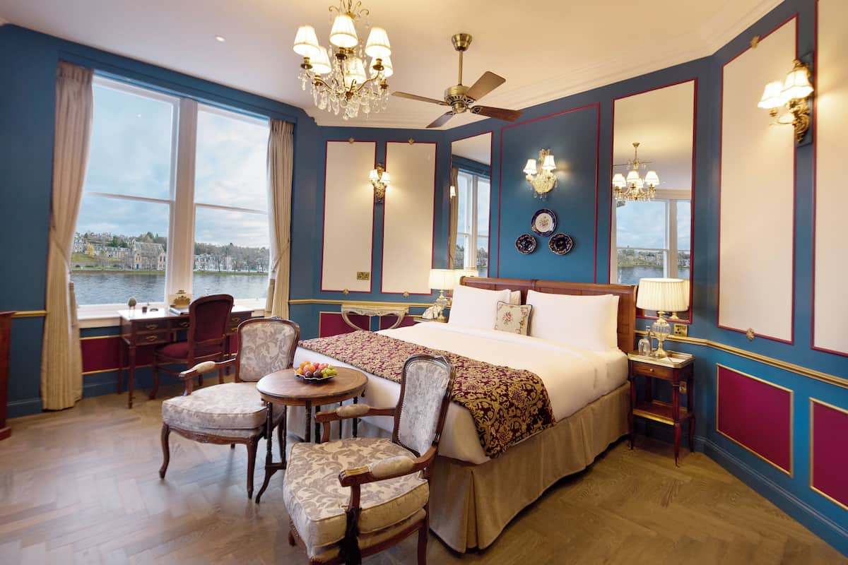 A hotel room with a large bed, a desk, two chairs, a small table with fruit, and a view of a body of water through large windows.