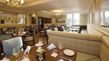 A dining area with multiple tables set for guests, featuring chandeliers and large windows.