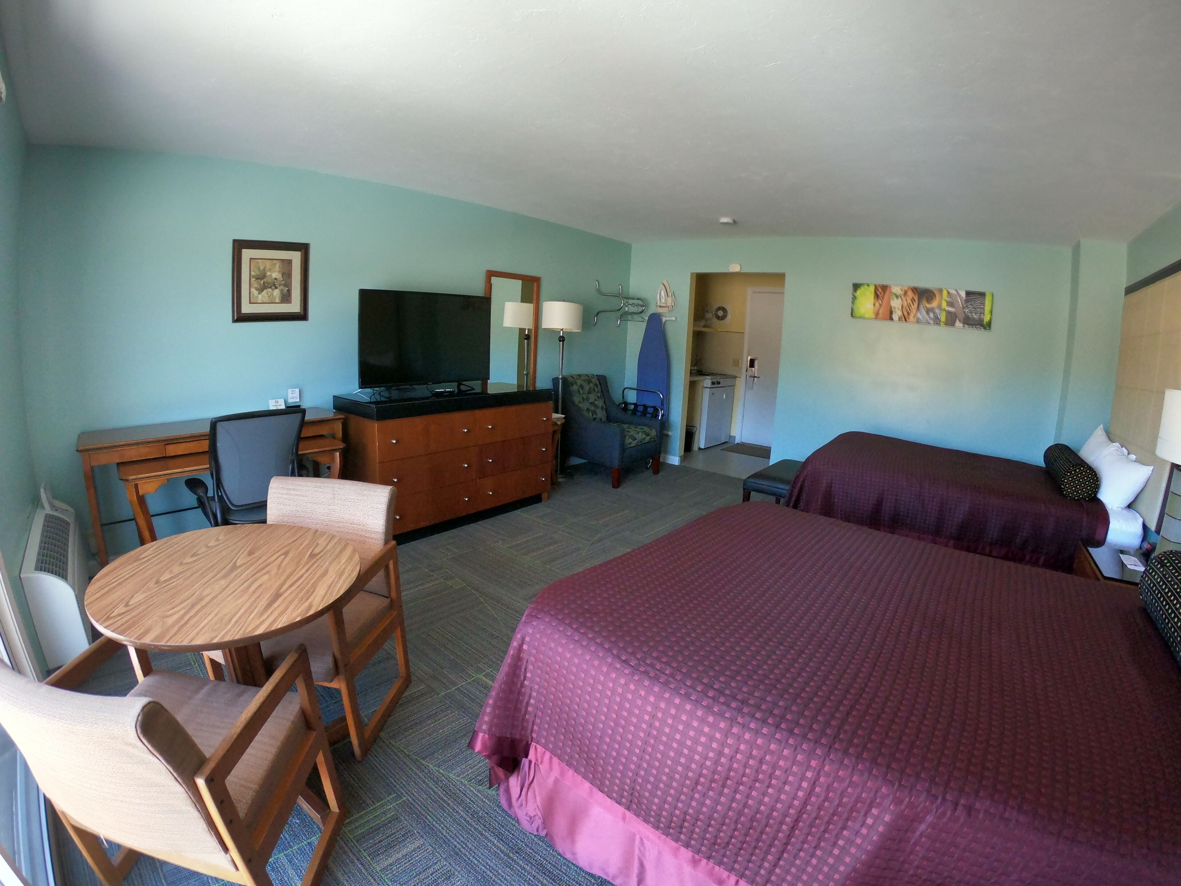 A hotel room with a bed, a desk with a chair, a television, a small kitchenette, and a bathroom.