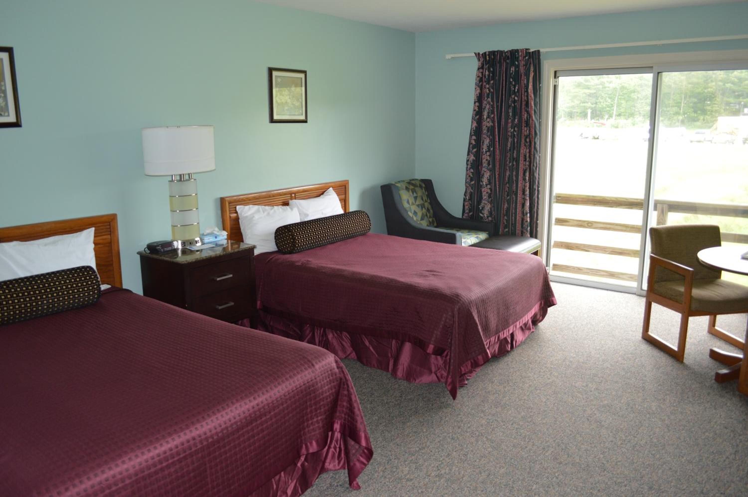 A hotel room with two beds, a chair, a table, and a view of the outdoors.