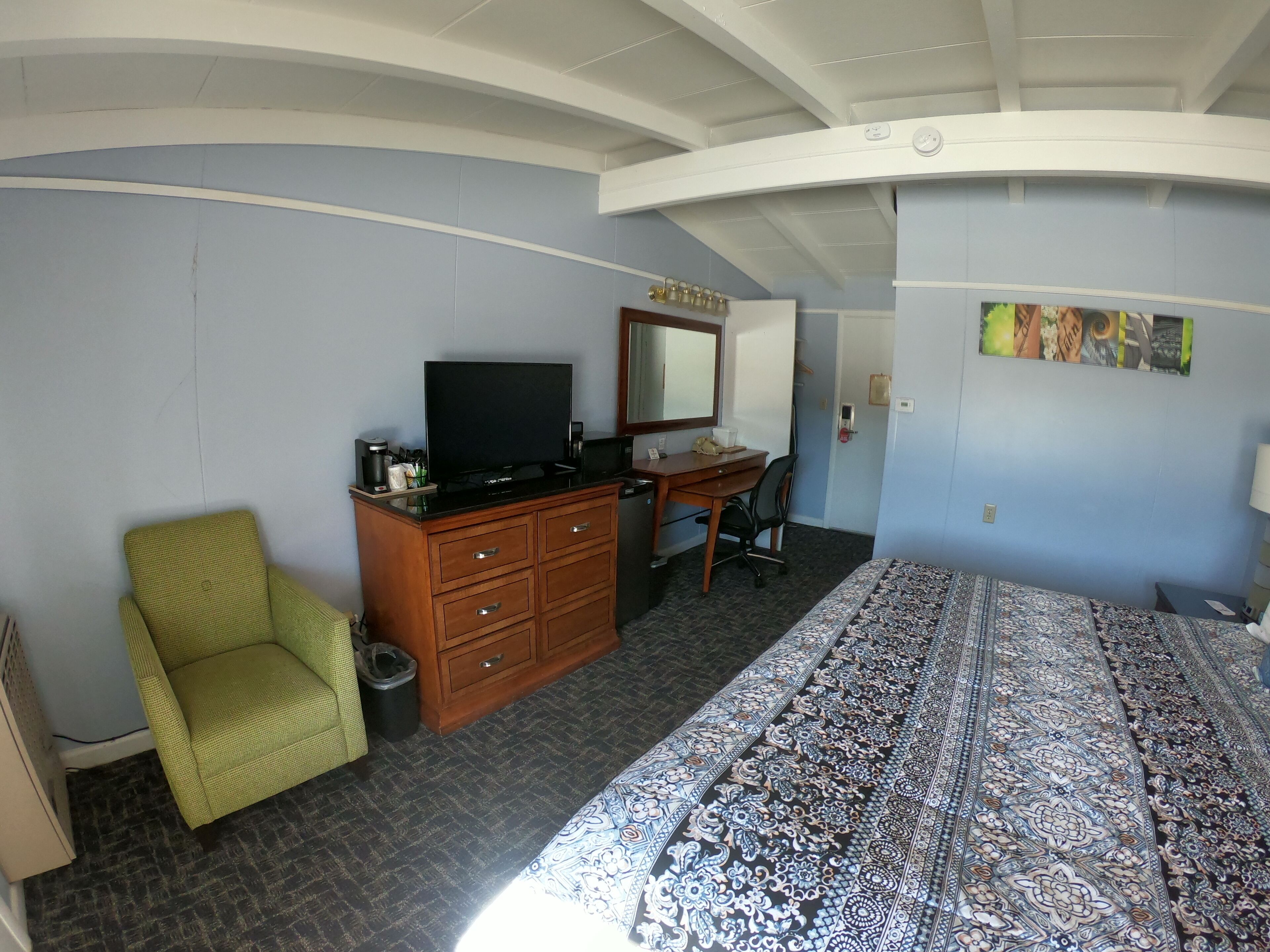 Una habitación de hotel con una cama, un escritorio con computadora, televisión, una silla y un espejo grande.