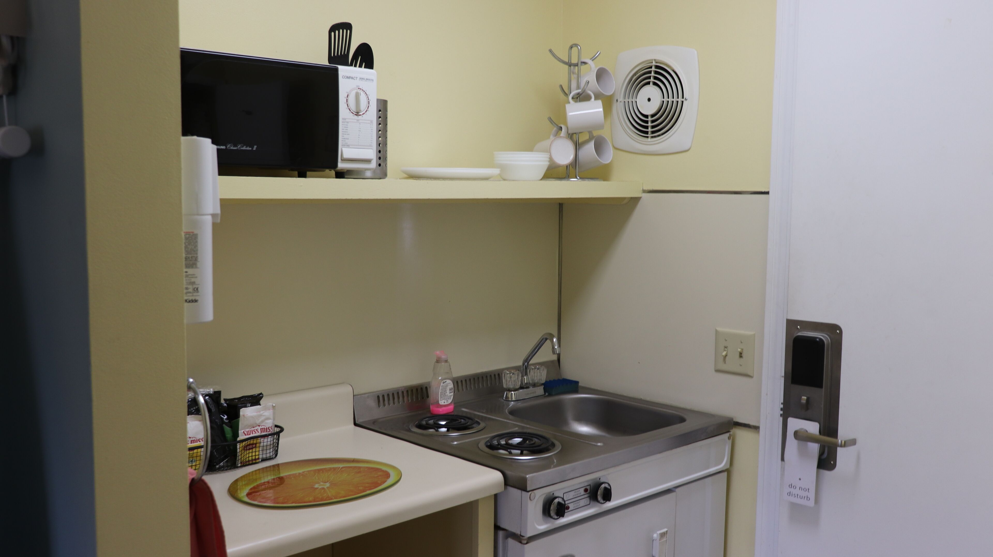 A compact kitchenette with a stove, sink, microwave, and a small fan.