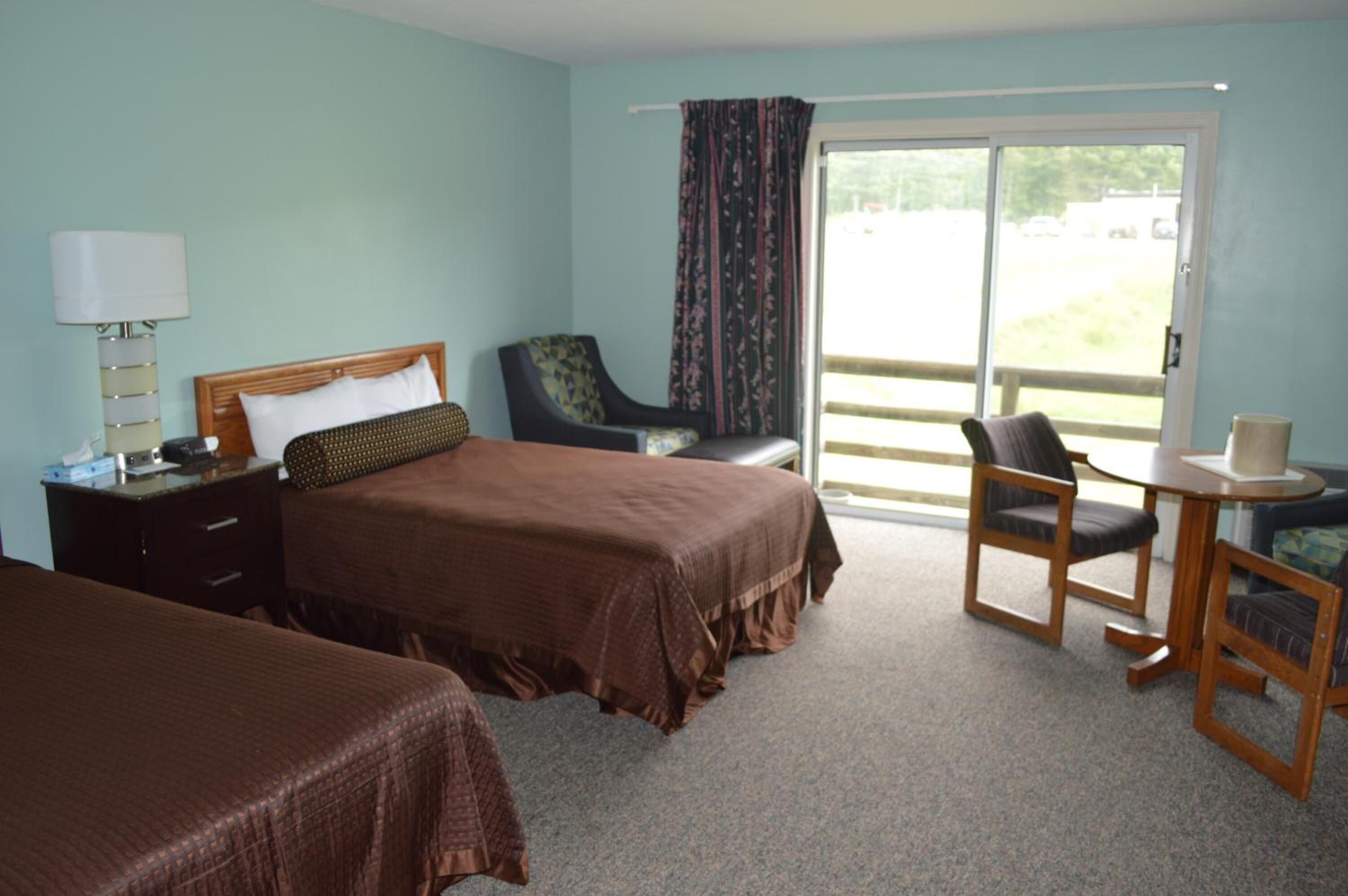 A hotel room with two beds, a chair, a table, and a view of the outdoors.