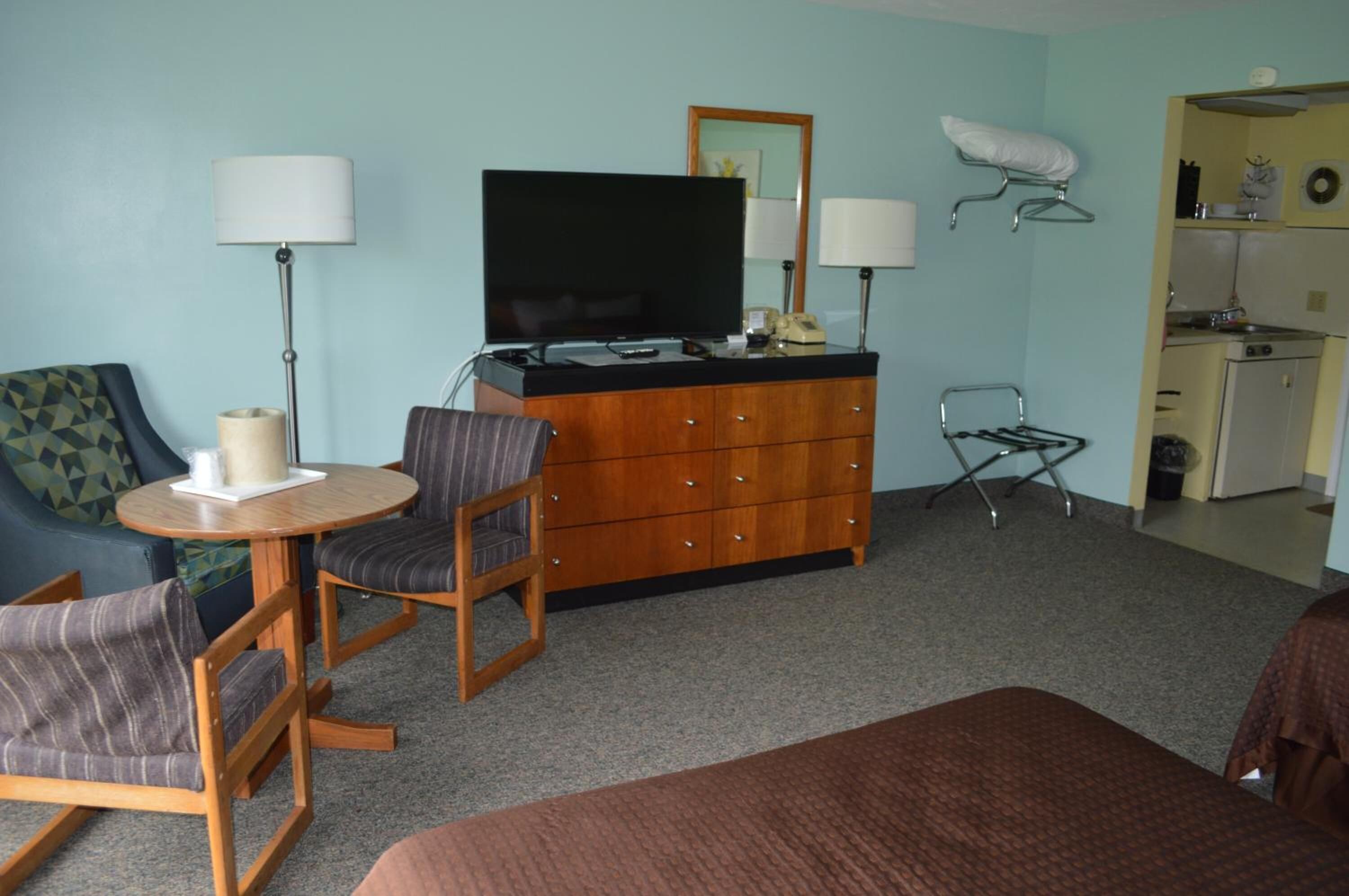 A hotel room with a bed, a wooden dresser, a television, two chairs, a small table, and a kitchenette.