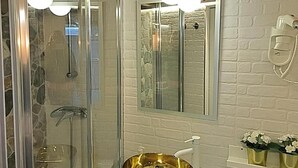Un baño con ducha envuelta en cristal, un lavamanos dorado y una pared de ladrillo blanco.
