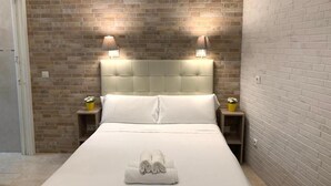 Hotelkamer met een bakstenen muur, een gestoffeerd hoofdeinde, twee bedlampen, een netjes opgemaakt bed met een gevouwen handdoek en een tafeltje met een bloevas.