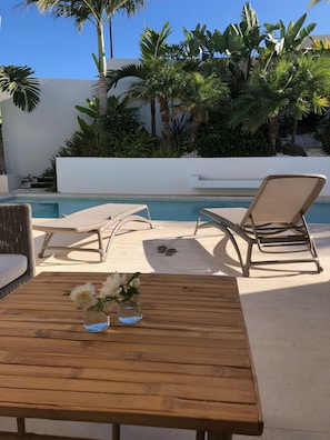 A poolside area with lounge chairs, a wooden table with a vase of flowers, and a clear blue pool.