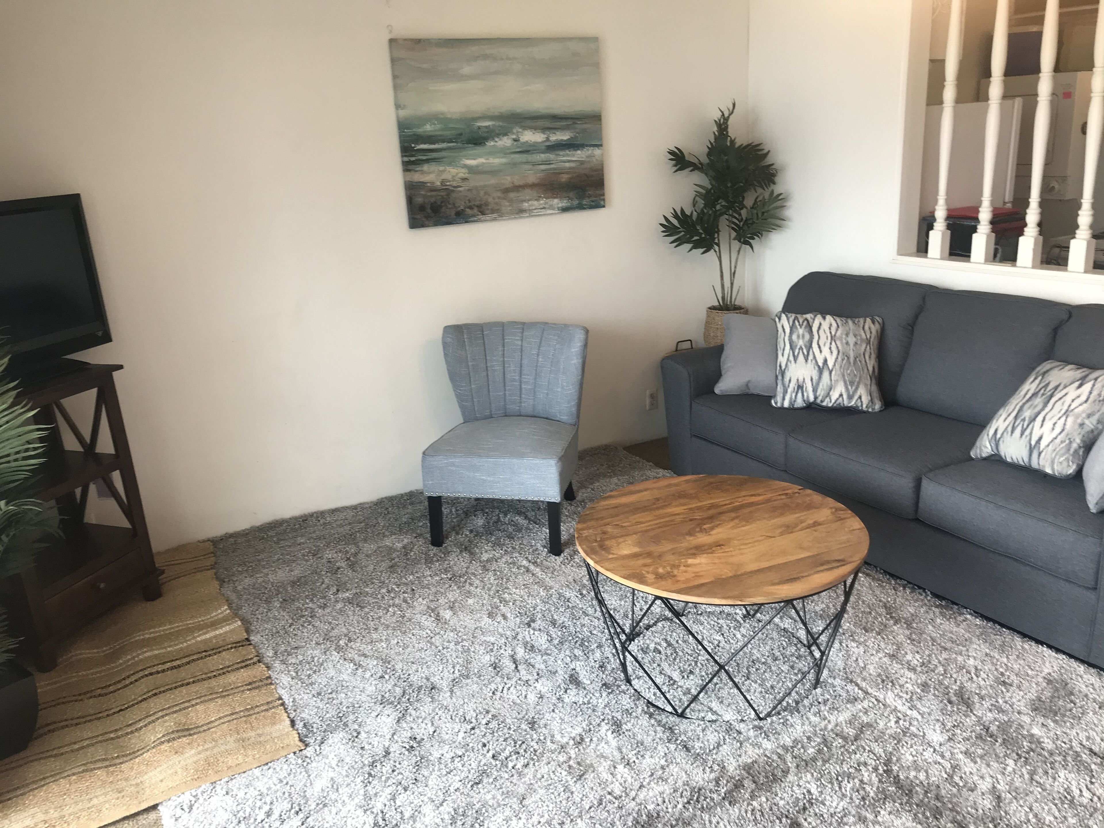Een woonkamer met een grijze bank, een houten salontafel, een grijze fauteuil, een schilderij aan de muur en een kamerplant.