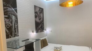 Un dormitorio con un escritorio de vidrio, una cama con colchón y almohadas blancas, y una lámpara fijada a la pared.