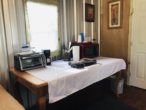 Un coin cuisine équipé d’un micro-ondes, d’un grille-pain et d’une machine à café, le tout posé sur une table, une fenêtre avec des rideaux et une porte ornée d’un tableau encadré.