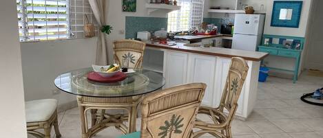 A kitchen with a dining area, featuring a round glass table, wicker chairs, and a small island with a sink.