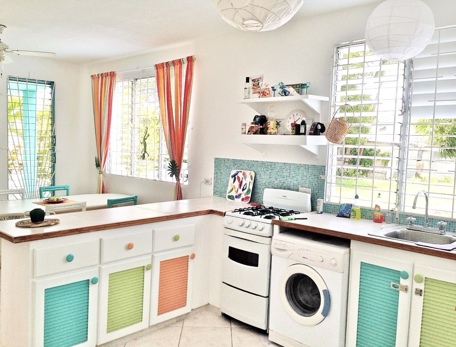 A kitchen with a gas stove, oven, and washing machine, a dining table with chairs, and a window with curtains.
