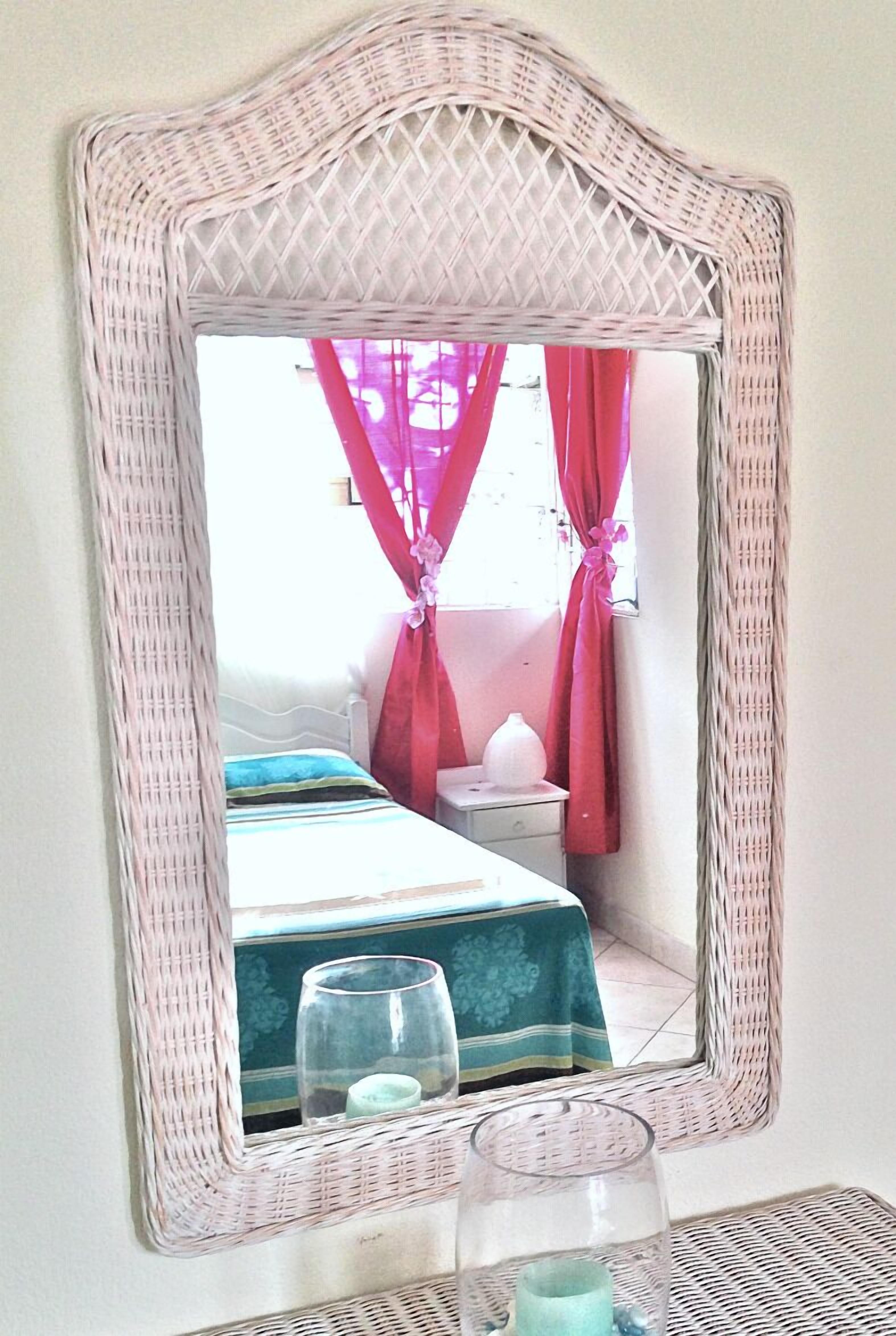 A mirror with a woven frame reflecting a room with a bed, a nightstand, and pink curtains.