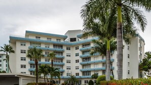 A multi-story hotel with a white facade and blue accents, featuring balconies, a paved driveway, and palm trees. - Beachfront efficiency studio with 1 bath (Marco Island)