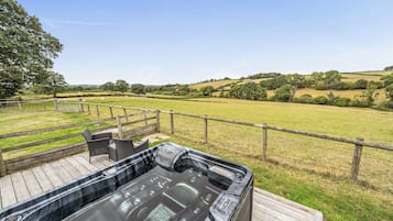 A hot tub on a wooden deck with a view of a grassy field and hills.