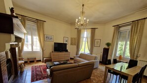 A living room with a sofa, a television, a fireplace, a chandelier, and a dining area. - 🏡 Spacious, air-conditioned 8-person gîte with swimming pool, between history, nature and the sea (Sommières)