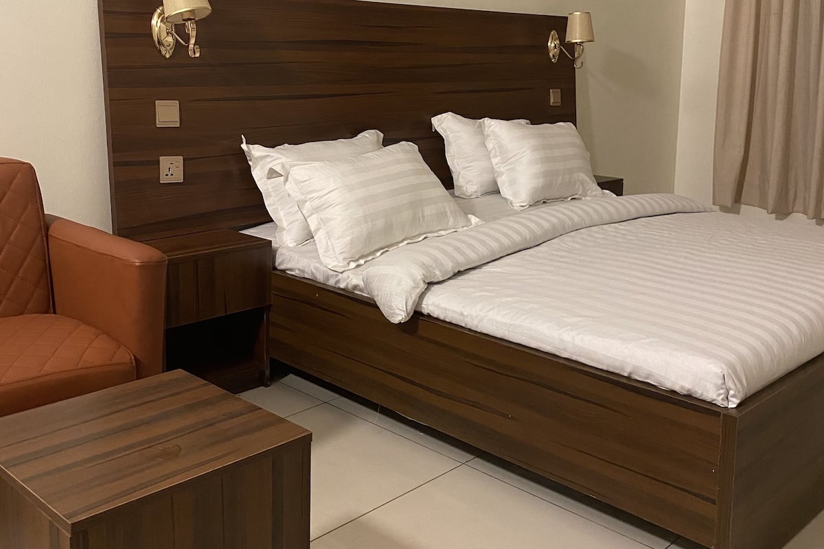 A bedroom with a wooden headboard, two bedside lamps, a bed with white bedding, a nightstand, a wooden side table, and a brown armchair.