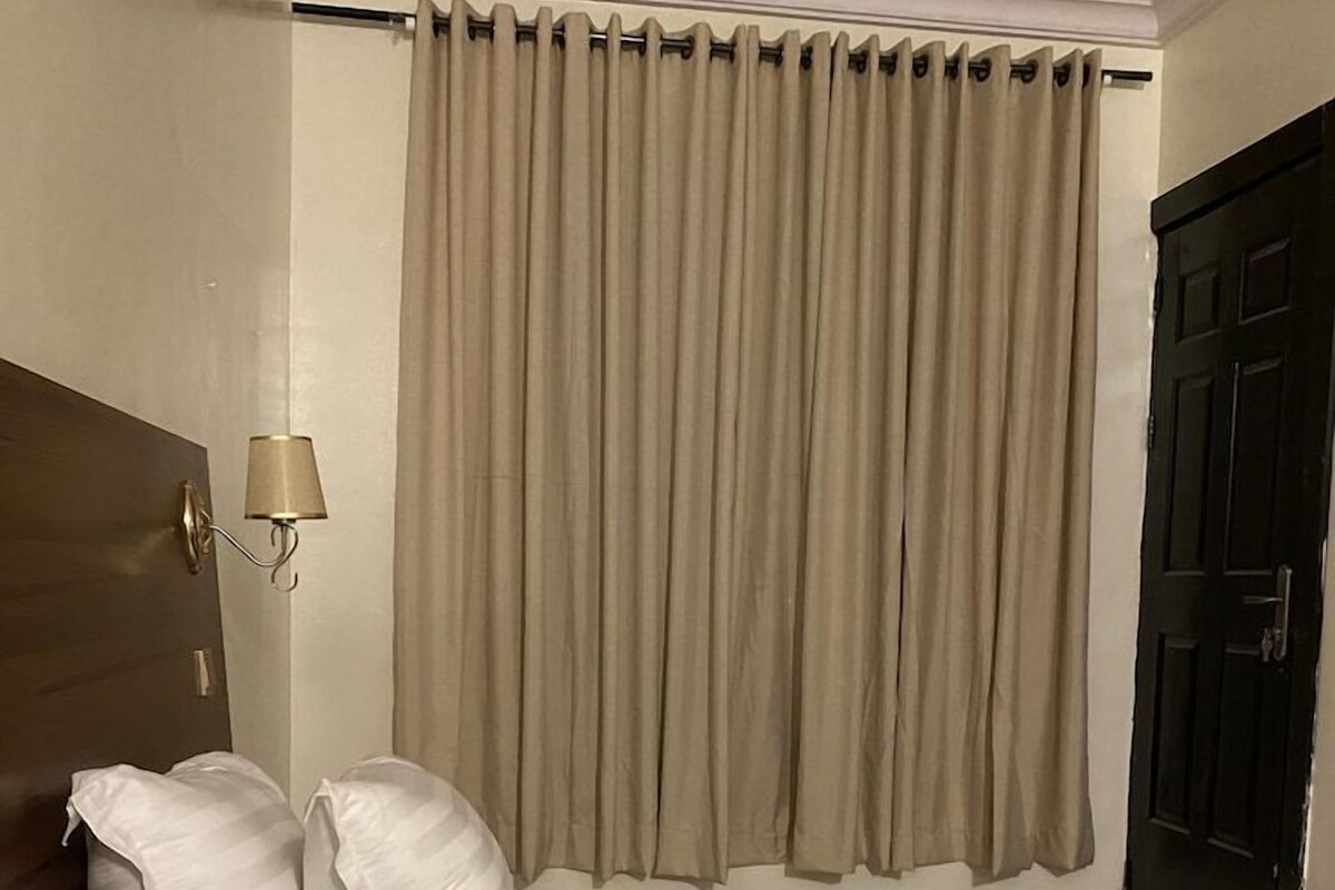 Habitació d'hotel amb dos llits, un capçal, una làmpada penjada a la paret i una finestra amb cortines.