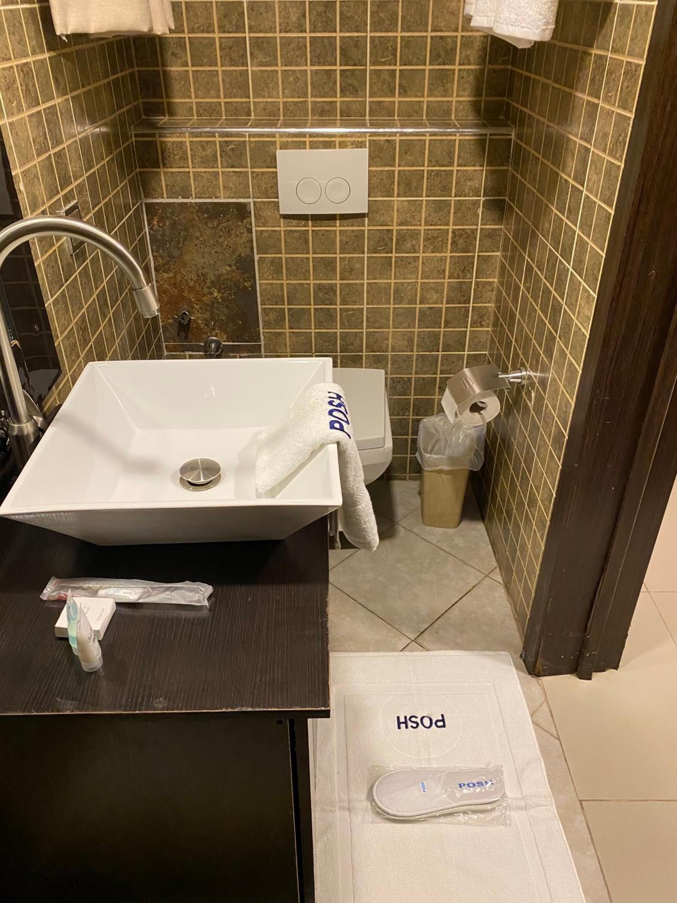 A bathroom with a white sink, a toilet, and a trash bin. There is a towel hanging on the wall and a package on the floor.
