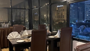 A dining area with a large table set for four, surrounded by dark brown chairs with metal frames. Large windows offer a view of the cityscape at night. - Posh Hotel And Suites (Lagos)