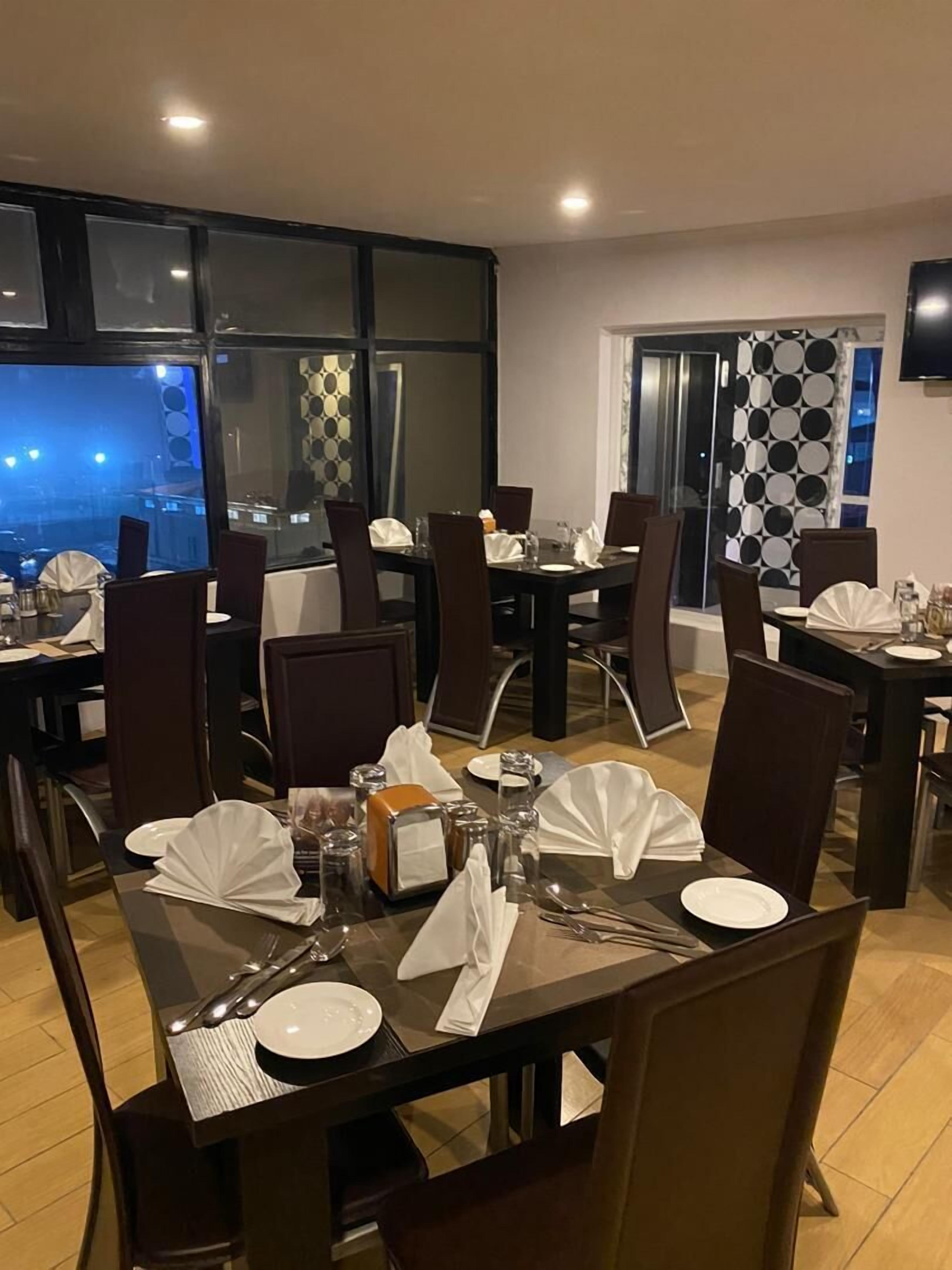 A dining area with tables set for four, each with white plates, cutlery, and napkins. The room has large windows with a view of the cityscape at night.