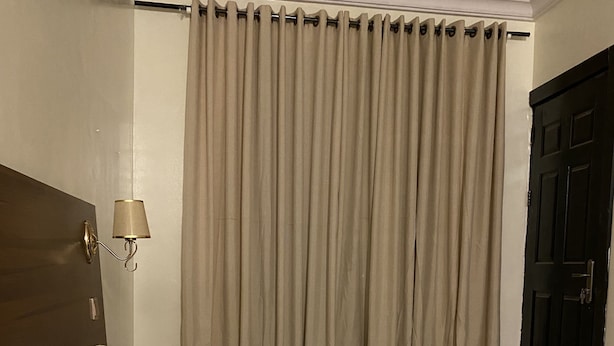 A hotel room with a large bed, two pillows, a headboard, a wall-mounted lamp, and a window with curtains. - Posh Hotel And Suites (Lagos)