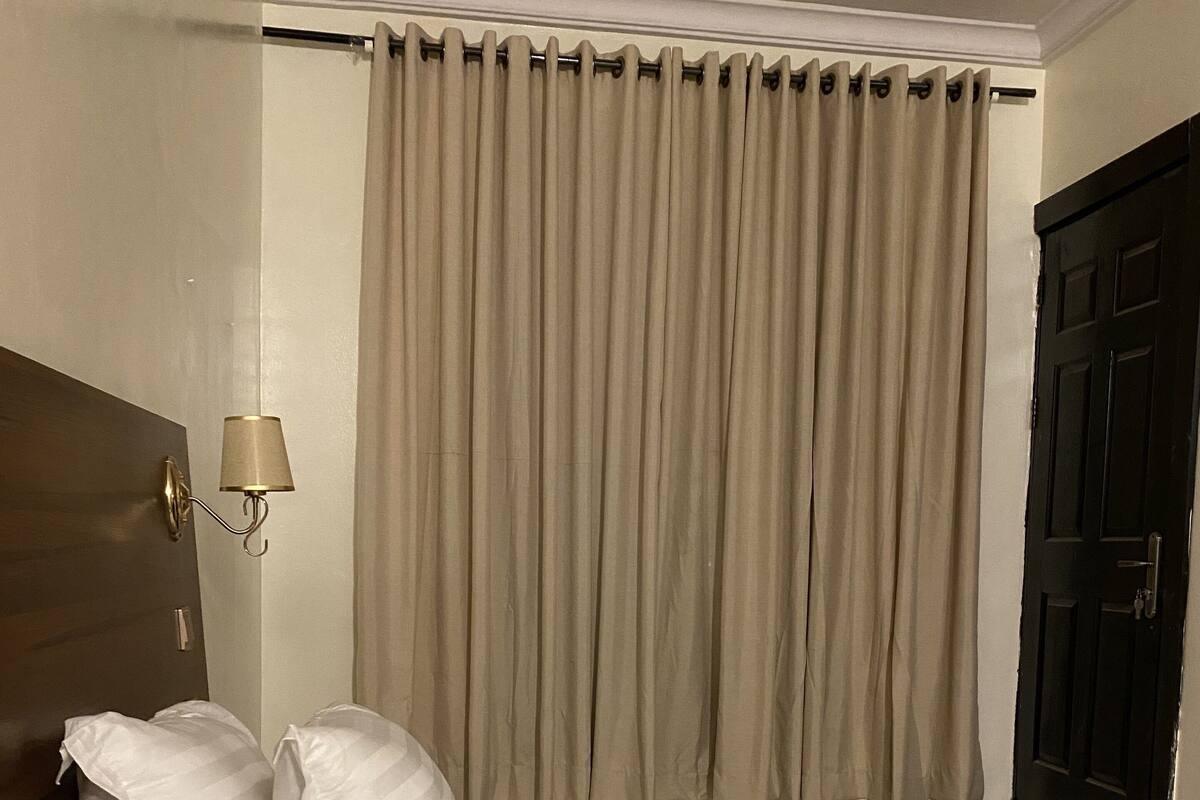 A hotel room with a large bed, two pillows, a headboard, a wall-mounted lamp, and a window with curtains.