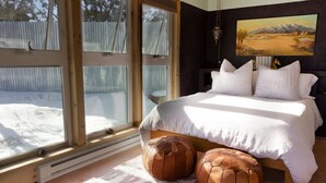 Un dormitorio con una cama grande, dos otomanas, un cuadro en la pared y vistas a un paisaje nevado a través de amplias ventanas.